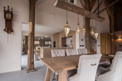 Sfeervolle leefkeuken en eetkamer in een woonboerderij met robuuste houten balken, landelijke houten keuken, grote eettafel en sfeervolle hanglampen boven de tafel.
